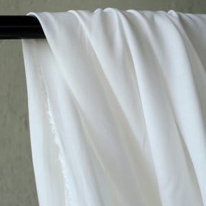 Pode incluir: Close-up de tecido branco drapeado sobre uma barra preta. O tecido tem uma textura macia e &eacute; ligeiramente transl&uacute;cido, com dobras vis&iacute;veis e uma borda crua. O fundo &eacute; verde suave.