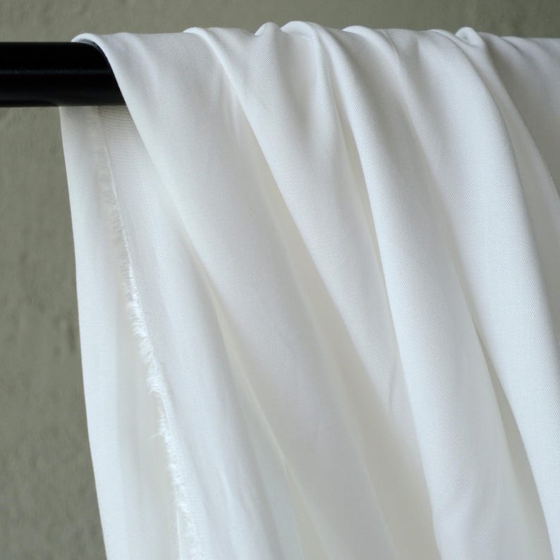 Pode incluir: Close-up de tecido branco drapeado sobre uma barra preta. O tecido tem uma textura macia e &eacute; ligeiramente transl&uacute;cido, com dobras vis&iacute;veis e uma borda crua. O fundo &eacute; verde suave.