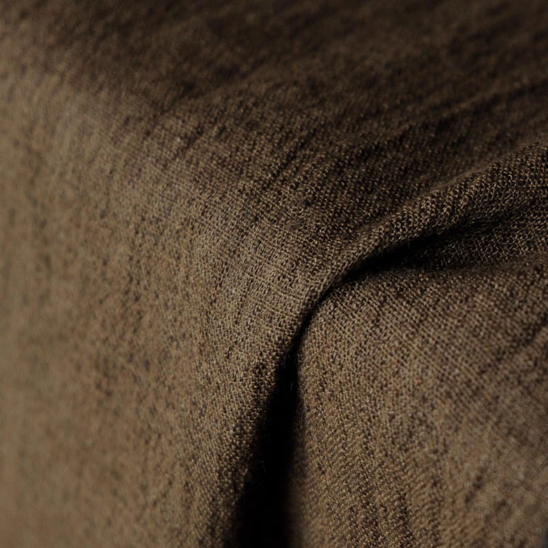 May include: Close-up of a textured, brown fabric with visible weave. The material is folded, creating shadows and depth. The color is a warm, earthy tone, suggesting a natural textile.