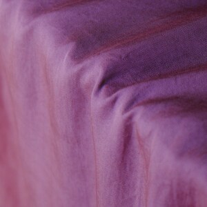 Pode incluir: Close-up de um tecido roxo texturizado com linhas diagonais vis&iacute;veis. O tecido tem um leve brilho, e as dobras criam sombras e varia&ccedil;&otilde;es de cor. O tom geral &eacute; um roxo suave e abafado.
