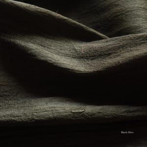 May include: Close-up of a dark olive green fabric with a textured, slightly wrinkled surface. The fabric has a subtle sheen and appears to be draped, with visible folds and shadows. The words "Black Olive" are in the bottom right corner.