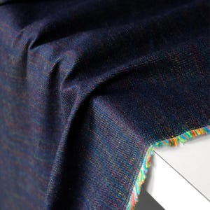 Op de afbeelding: Close-up van een donkerblauwe stof met een getextureerde weefsel. De stof heeft subtiele regenboogkleurige draden die er doorheen zijn geweven, waardoor een glinsterend effect ontstaat. De rand van de stof is afgewerkt met een kleurrijke franje.