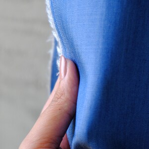 May include: Close-up of a piece of blue fabric with a frayed edge. The fabric is a solid, medium shade of blue. The texture of the fabric is smooth and appears to be soft. The edge of the fabric is held by a finger.