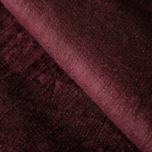 May include: Close-up of a folded piece of burgundy fabric. The fabric has a soft, textured appearance with visible fibers and subtle variations in color. The folds create shadows and depth, highlighting the material's texture.