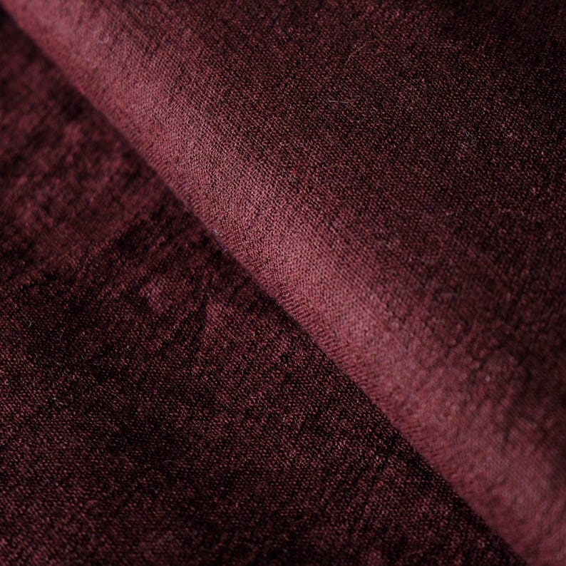 May include: Close-up of a folded piece of burgundy fabric. The fabric has a soft, textured appearance with visible fibers and subtle variations in color. The folds create shadows and depth, highlighting the material's texture.