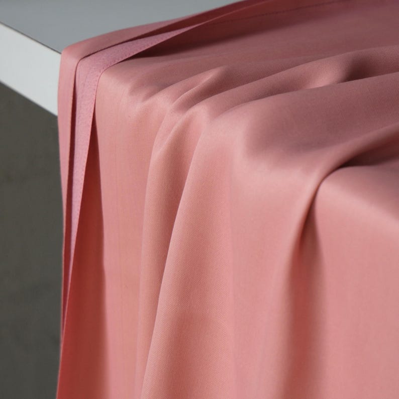 Puede incluir: Primer plano de una tela drapeada en un suave color rosa apagado. La tela tiene una textura suave y sedosa con pliegues y sombras sutiles, creando una sensaci&oacute;n de profundidad y movimiento. El borde de la tela est&aacute; doblado, agregando un efecto en capas.