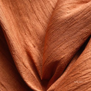May include: Close-up of a folded fabric in a warm toffee brown color. The textile has a textured appearance with visible woven threads and subtle variations in tone. The fabric is draped, creating soft folds and shadows. The words "Toffee Brown" are visible.