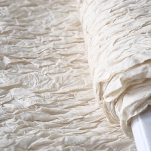 May include: Close-up of a roll of off-white fabric with a textured, ruffled surface. The fabric appears to be made of a soft, natural material, possibly linen or cotton. The texture creates a sense of depth and visual interest.