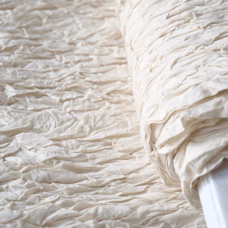 May include: Close-up of a roll of off-white fabric with a textured, ruffled surface. The fabric appears to be made of a soft, natural material, possibly linen or cotton. The texture creates a sense of depth and visual interest.