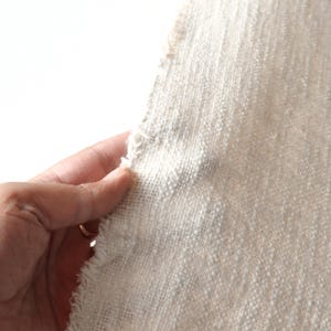May include: Close-up of a piece of off-white fabric with a textured weave. The fabric has a raw edge and is slightly frayed.