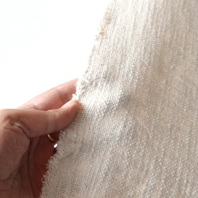 May include: Close-up of a piece of off-white fabric with a textured weave. The fabric has a raw edge and is slightly frayed.