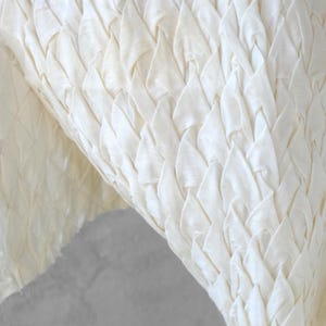 May include: Close-up of a cream-colored fabric with a woven, textured pattern. The fabric's design features a series of overlapping, triangular shapes, creating a layered effect. The material appears soft and pliable, with subtle variations in tone.
