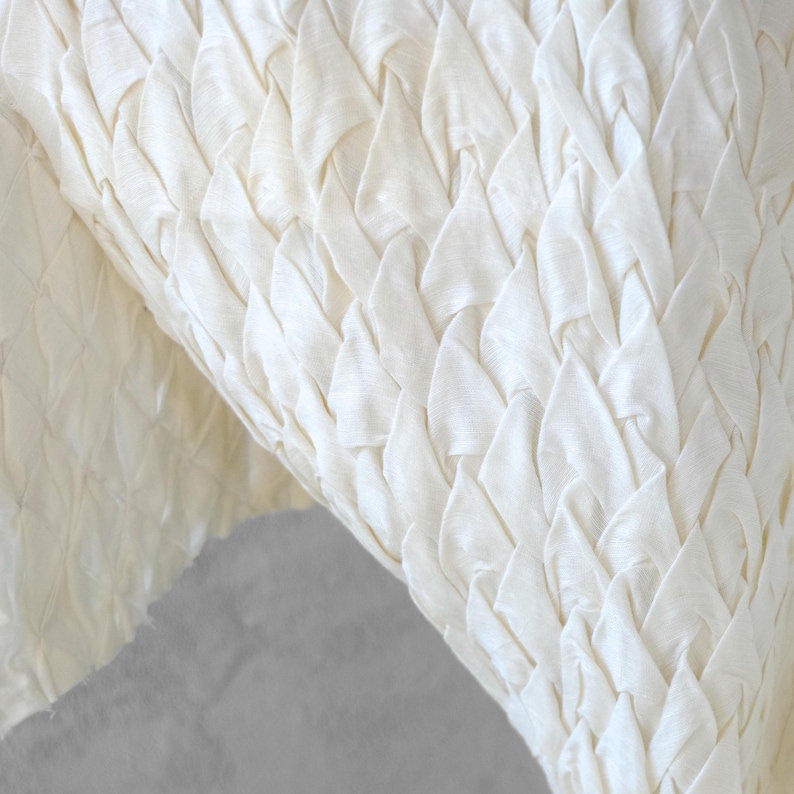 May include: Close-up of a cream-colored fabric with a woven, textured pattern. The fabric's design features a series of overlapping, triangular shapes, creating a layered effect. The material appears soft and pliable, with subtle variations in tone.