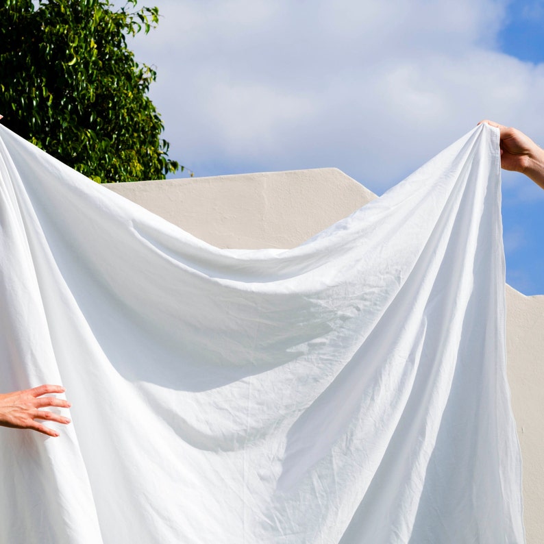 May include: A white sheet of fabric is held up by two hands against a white wall and a blue sky. The fabric is draped over the wall and the sky, creating a soft, flowing shape.
