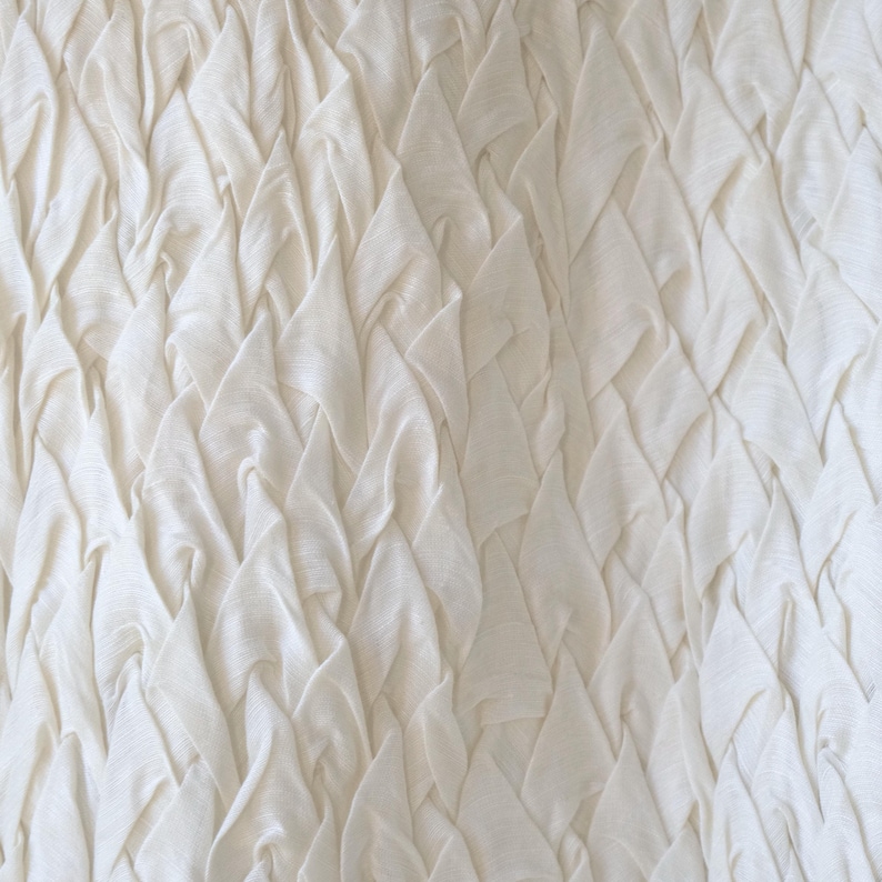 May include: Close-up of a textured, off-white fabric. The fabric is intricately folded and layered, creating a pattern of raised, triangular shapes. The folds and shadows give the fabric a three-dimensional appearance.