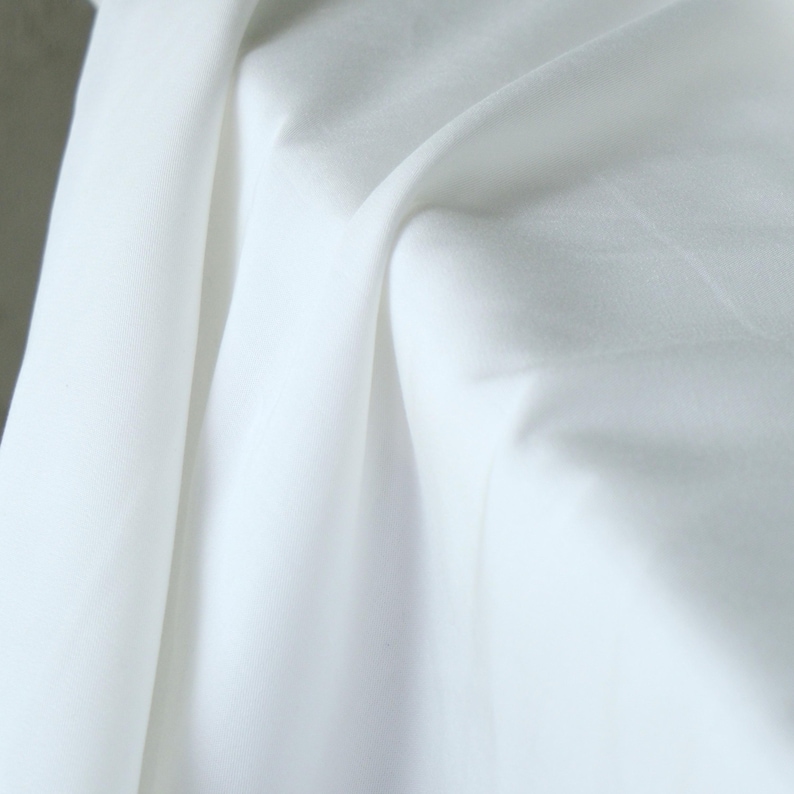 Pode incluir: Close-up de tecido branco com dobras suaves e sombras. O tecido parece liso e leve, possivelmente algod&atilde;o ou um material semelhante. A imagem destaca a textura e o caimento do tecido.