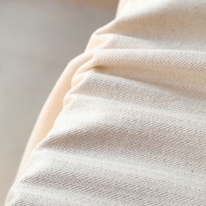 May include: Close-up of a cream-colored fabric with a herringbone weave pattern. The fabric is tightly woven and appears soft, with subtle variations in color and texture. The folds and shadows create depth and visual interest.