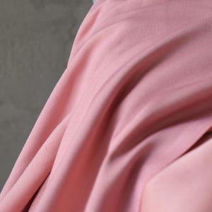 Puede incluir: Primer plano de una tela rosa claro y fluida. La tela tiene sutiles l&iacute;neas verticales y una textura suave y lisa. Los pliegues y drapeados crean una sensaci&oacute;n de movimiento y profundidad, con diferentes tonos de rosa.