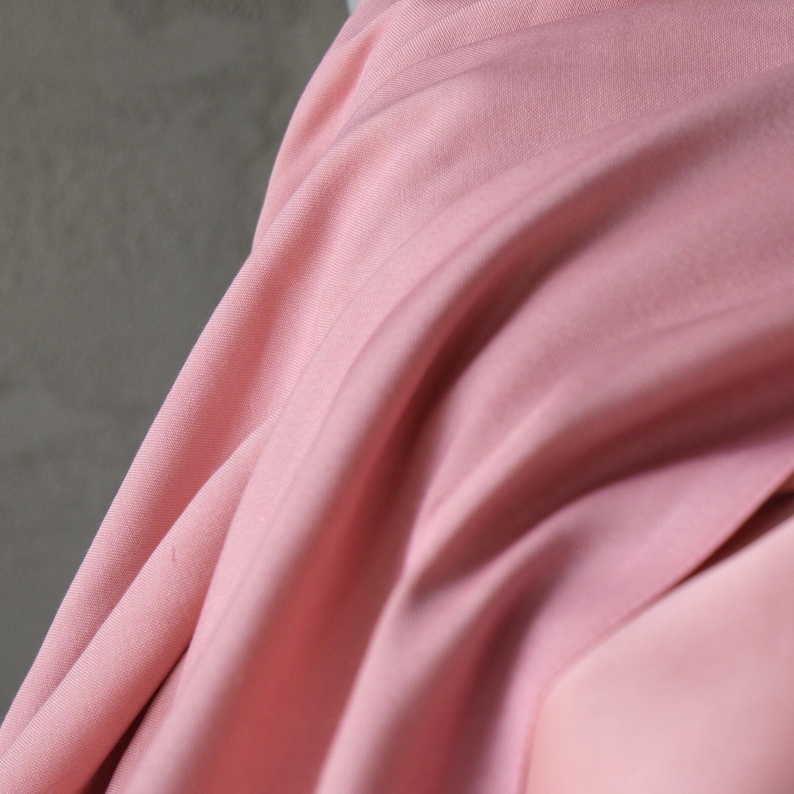 Puede incluir: Primer plano de una tela rosa claro y fluida. La tela tiene sutiles l&iacute;neas verticales y una textura suave y lisa. Los pliegues y drapeados crean una sensaci&oacute;n de movimiento y profundidad, con diferentes tonos de rosa.