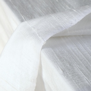 May include: Close-up of white fabric with a textured, ribbed pattern. The fabric is folded over, revealing the underside and the stitched edge. The lighting creates shadows and highlights, emphasizing the texture and the weave of the material.