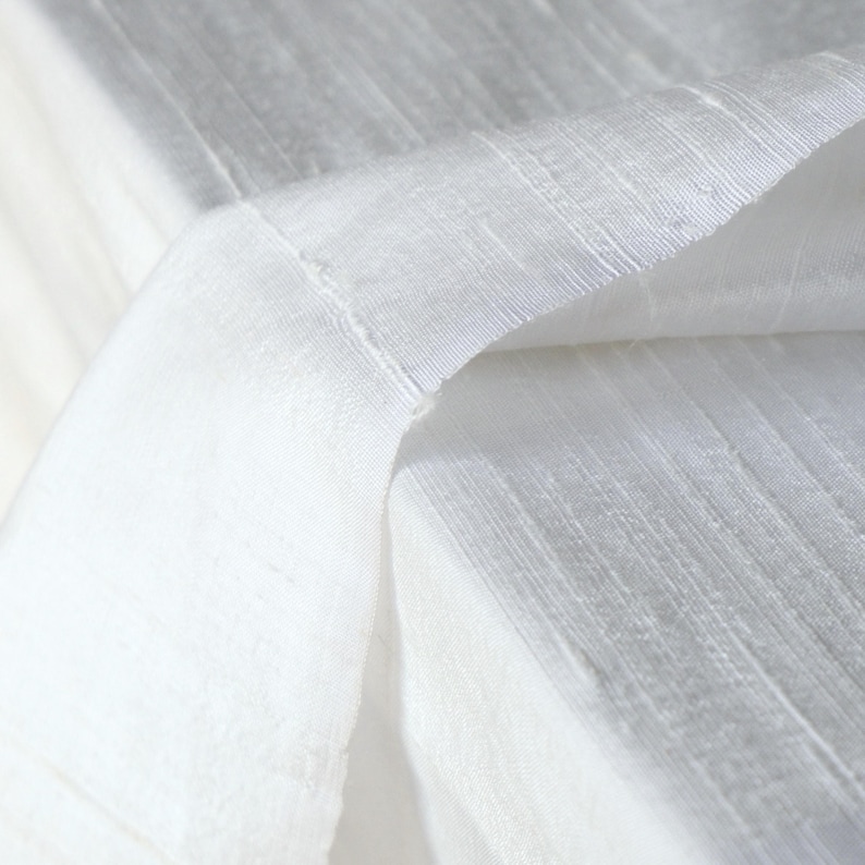 May include: Close-up of white fabric with a textured, ribbed pattern. The fabric is folded over, revealing the underside and the stitched edge. The lighting creates shadows and highlights, emphasizing the texture and the weave of the material.