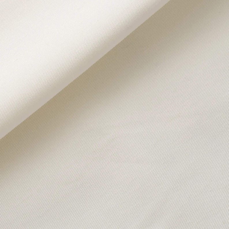 K&ouml;nnte beinhalten: Eine Nahaufnahme von einfarbigem wei&szlig;em Stoff mit einem ausgepr&auml;gten diagonalen K&ouml;perbindungsmuster. Das Material ist an der oberen linken Ecke umgeschlagen und zeigt die Textur und die gleichm&auml;&szlig;ige cremewei&szlig;e Farbe der Textiloberfl&auml;che.