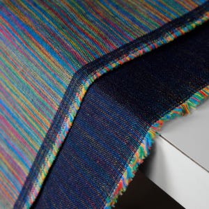 Op de afbeelding: Close-up van een levendig textiel met een streepjespatroon in tinten blauw, rood, geel en groen. De stof is dubbelgevouwen, waardoor een donkerblauwe onderkant zichtbaar is. De randen zijn afgewerkt met kleurrijke franjes.