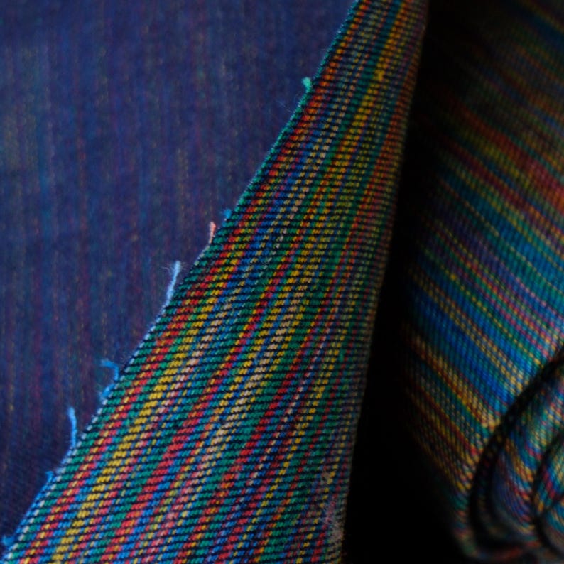 Op de afbeelding: Close-up van een geweven stof met een levendig streepjespatroon. De stof heeft een diepblauwe basis met diagonale strepen in rood, geel, groen en blauw. De randen vertonen losse draden, wat duidt op een stuk materiaal dat is gesneden.