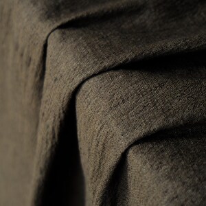 May include: Close-up of a textured, dark brown fabric with visible weave details. The fabric is draped, creating folds and shadows that highlight its texture and depth. The material appears to be a heavy linen or similar textile.