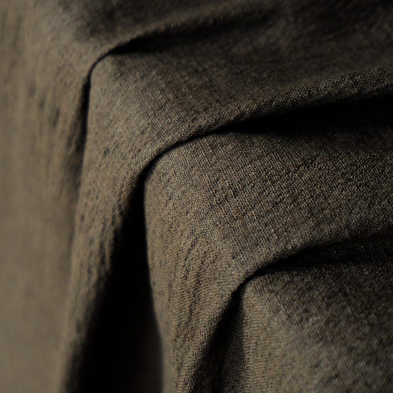 May include: Close-up of a textured, dark brown fabric with visible weave details. The fabric is draped, creating folds and shadows that highlight its texture and depth. The material appears to be a heavy linen or similar textile.