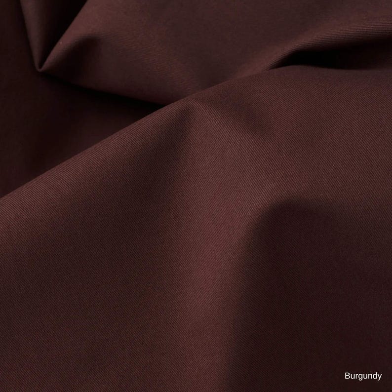K&ouml;nnte beinhalten: Ein einfarbiger, dunkelburgunderroter Stoff mit einer glatten, matten Textur und einer dichten, gleichm&auml;&szlig;igen Webart. Das Material ist in weichen, flie&szlig;enden Falten drapiert, die tiefe Schatten und Lichter auf der Oberfl&auml;che erzeugen, mit dem Wort Burgundy in der Ecke.