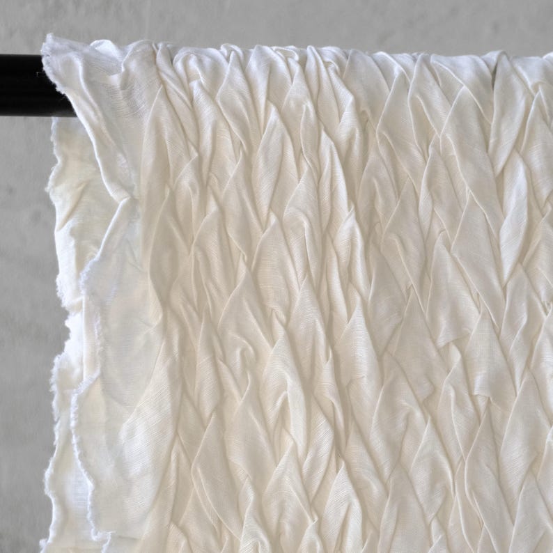 May include: Off-white fabric with a textured, woven pattern. The fabric is draped over a black rod, with the edges of the fabric showing a raw, unfinished edge. The fabric appears to be lightweight and airy.