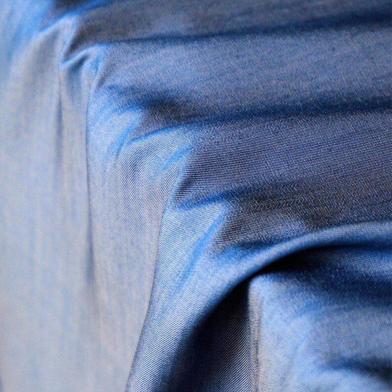 May include: Close-up of blue fabric with a subtle gradient effect. The fabric has a textured appearance, with visible woven threads. The folds and shadows create depth and dimension, highlighting the material's drape and texture. The colour transitions from a lighter blue to a darker shade.