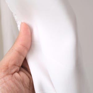 Puede incluir: Primer plano de tela blanca, sostenida por una mano. La tela parece ser un material ligero, posiblemente una tela transparente o semitransparente. Los bordes de la tela son visibles, mostrando un dobladillo terminado. El fondo es de un tono suave y neutro.