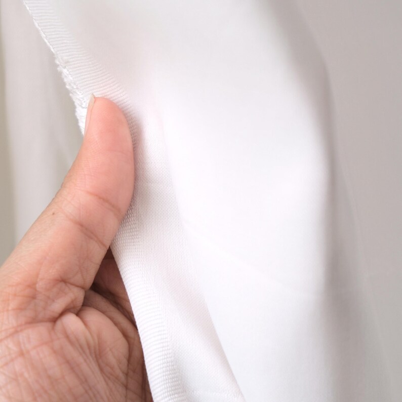 Puede incluir: Primer plano de tela blanca, sostenida por una mano. La tela parece ser un material ligero, posiblemente una tela transparente o semitransparente. Los bordes de la tela son visibles, mostrando un dobladillo terminado. El fondo es de un tono suave y neutro.