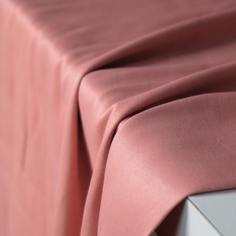 Puede incluir: Primer plano de una tela rosa empolvado drapeada sobre una superficie. La tela tiene una textura suave y est&aacute; doblada en suaves ondas fluidas. El color es un rosa apagado, con sutiles variaciones de tono debido a los pliegues y la iluminaci&oacute;n.