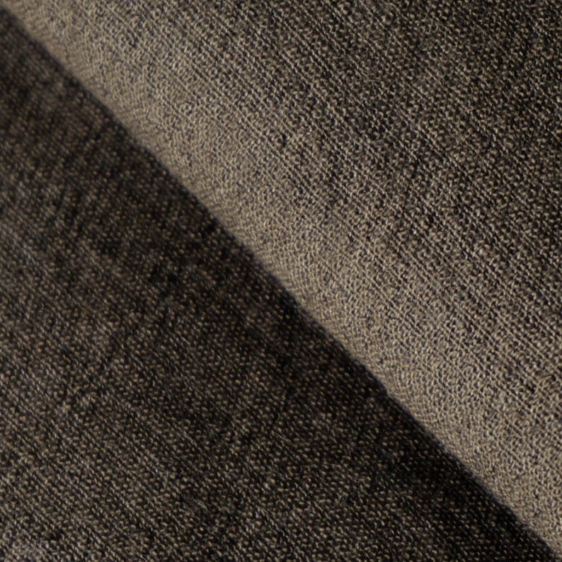 May include: Close-up of a folded section of dark brown fabric. The fabric has a textured, woven appearance, with visible threads and a slightly rough surface. The color varies from dark brown to a lighter, muted brown.