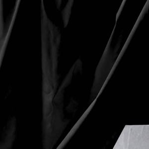 May include: A solid black fabric with a smooth, slightly reflective surface texture. The material is draped in deep, vertical folds that create varying shadows and highlights across the dark surface, emphasizing the depth and movement of the textile.