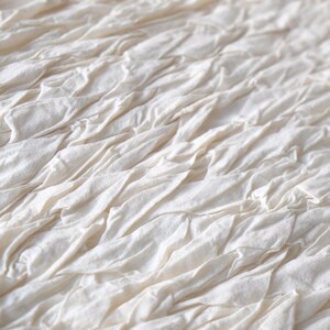 May include: Close-up of a textured, off-white fabric. The fabric is composed of layered, gathered strips, creating a rippled effect. The material appears soft and has a natural, organic aesthetic. The overall look is neutral and versatile.
