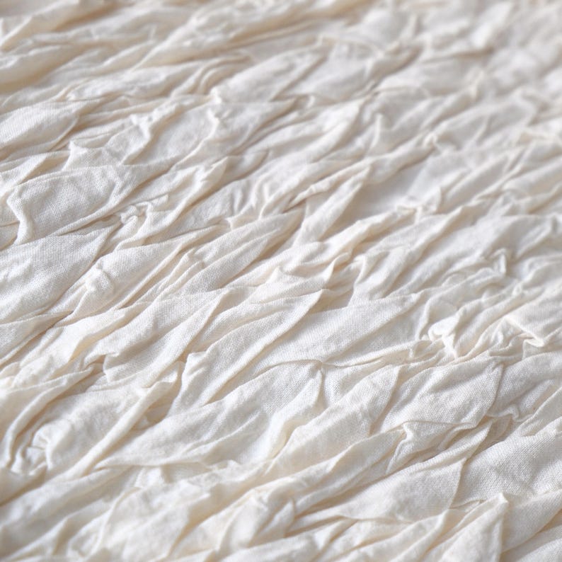 May include: Close-up of a textured, off-white fabric. The fabric is composed of layered, gathered strips, creating a rippled effect. The material appears soft and has a natural, organic aesthetic. The overall look is neutral and versatile.