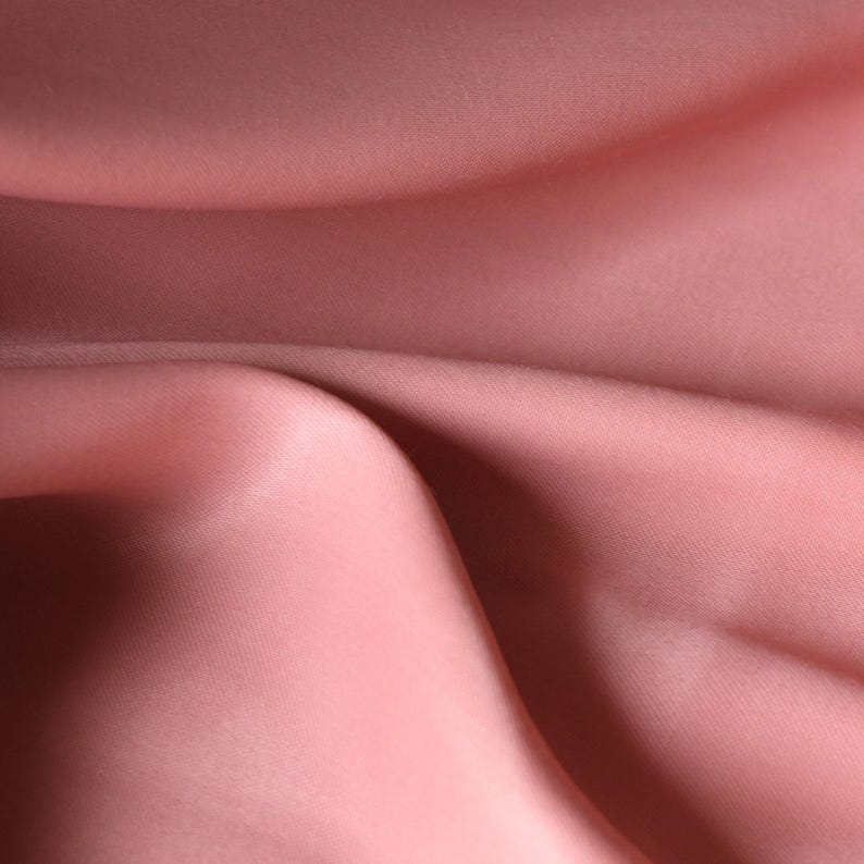 Puede incluir: Primer plano de una tela rosa rubor, posiblemente seda o sat&eacute;n. El material est&aacute; drapeado, con pliegues suaves y sombras que crean una sensaci&oacute;n de movimiento y textura. El efecto general es suave y elegante.