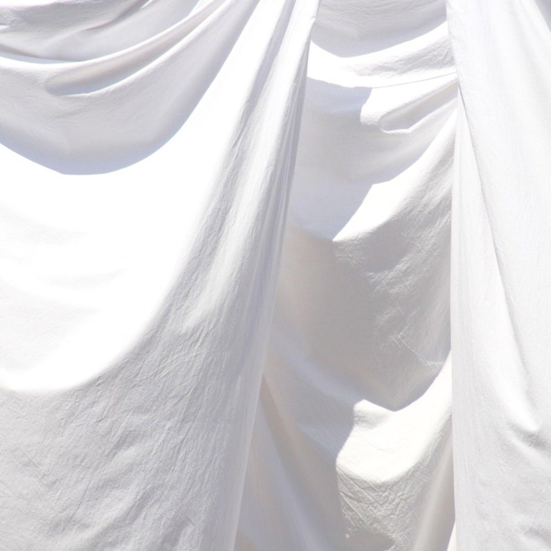 May include: White fabric sheets hanging on a clothesline, creating soft folds and shadows. The fabric is smooth and appears to be freshly laundered.