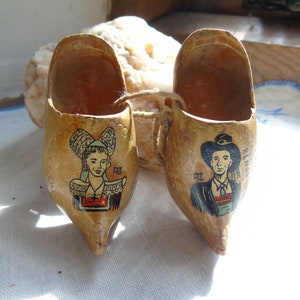 Könnte beinhalten: Ein Paar Holzschuhe mit bemalten Bildern einer Frau und eines Mannes in traditioneller Kleidung. Die Clogs sind braun und haben ein hellbraunes Innenleben.