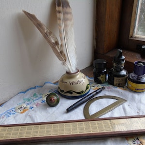 May include: A vintage wooden slide rule with a brass protractor, a black pen, a small wooden top, and three ink bottles with feathers on a white lace tablecloth. The ink bottles are labeled "Whitley" and "Waterman's Ideal."