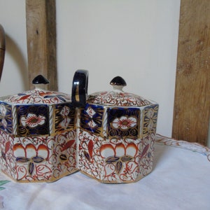 May include: A pair of octagonal porcelain tea caddies with lids. The caddies are decorated with a floral pattern in shades of red, blue, and gold. The caddies are joined together by a black handle.