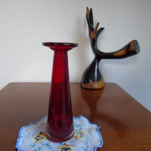 May include: A dark red glass vase with a flared top sits on a white doily with blue trim and floral embroidery. The vase is on a wooden table with a dark wood sculpture of a deer head in the background.