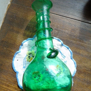 Peut inclure: Un vase en verre vert avec un corps bulbeux et un col torsadé. Le vase est posé sur un napperon blanc à broderie florale.