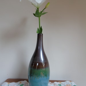 May include: A decorative vase holding a white lily. The vase has a glossy finish and a colour gradient, with brown at the top, green in the middle, and turquoise at the base. The lily has white petals and a green stem.