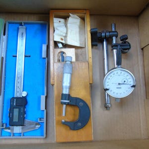 May include: A set of three measuring tools in a wooden box. The tools include a digital caliper, a micrometer, and a dial gauge. The caliper is silver with a black digital display. The micrometer is silver with a black handle. The dial gauge is silver with a black dial and a black pointer.