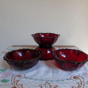 Può includere: Tre ciotole di vetro rosso con bordo smerlato. Le ciotole sono disposte a forma di piramide su una tovaglia bianca con ricamo floreale.